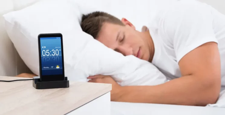Young Man Sleeping On Bed With Alarm On Mobile Phone Display